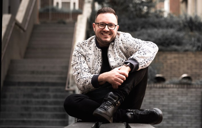 Singer, songwriter Matt Wright sitting on some stone steps, smiling, wearing black jeans and a black and white jacket.