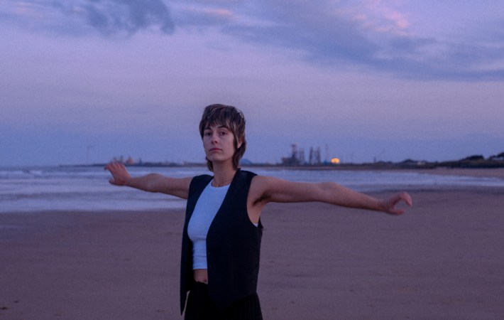 Sunset, and Newcastle-based indie-rock artist Melanie Baker standing at a beach, arms wide to the sides, looking with interest and a bit of sceptism into the camera.